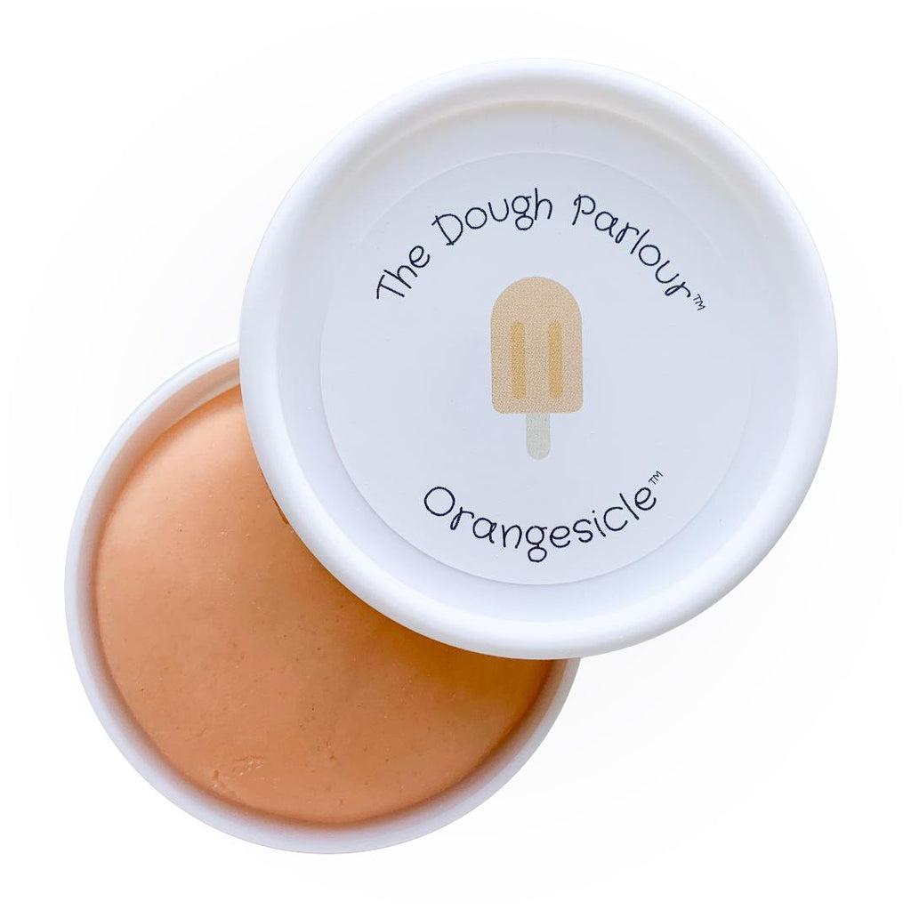 White background with Orangesicle Play Dough by Dough Parlour, open slightly to show colour. Play dough comes in a white container, dough is orange, and lid has a sticker that says "Orangesicle".