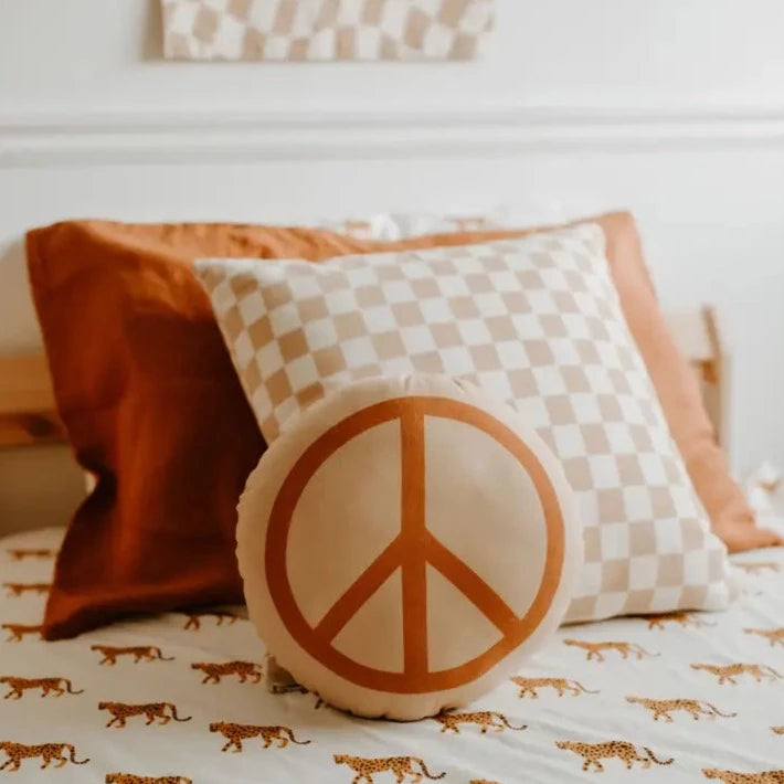 Peace Pillow in Peach by Imani Collective, with rust and check pillow behind it on a bed, with a sheet with cheetahs. 