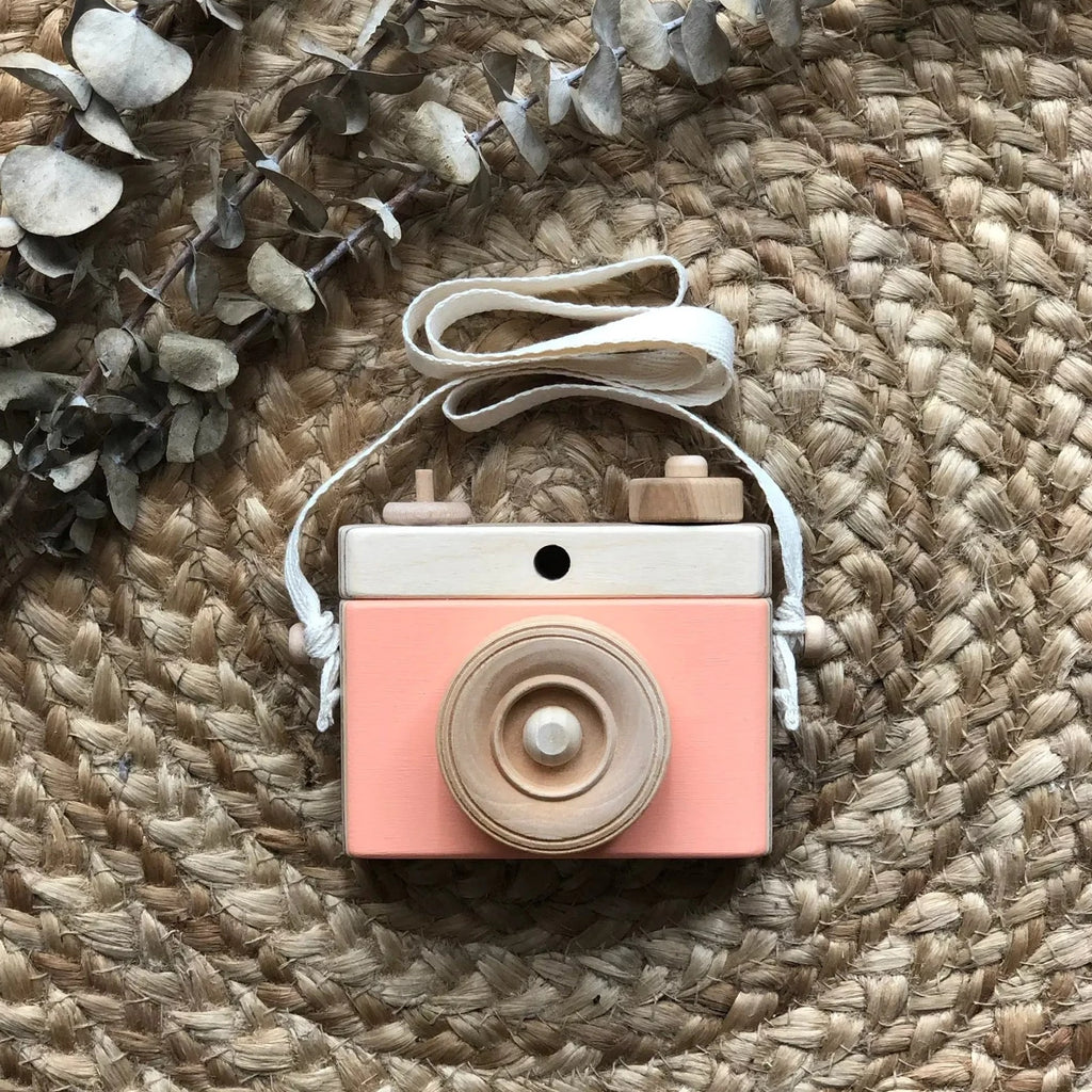 Rattan background with a Wooden Camera in Peach by Little Rose & Co in the centre. Camera is wood with peach accents, and a canvas strap.