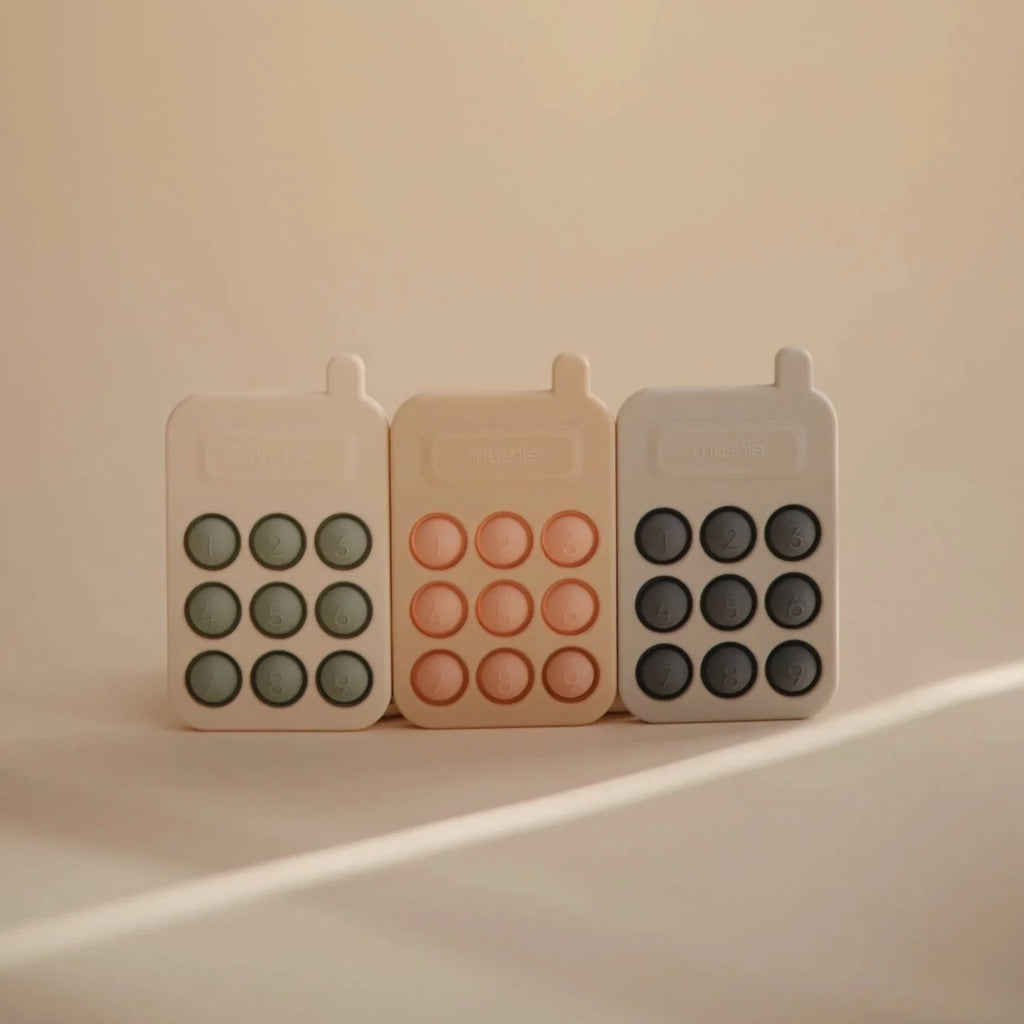 Beige background with the 3 Phone Press Toys by Mushie lined up together.