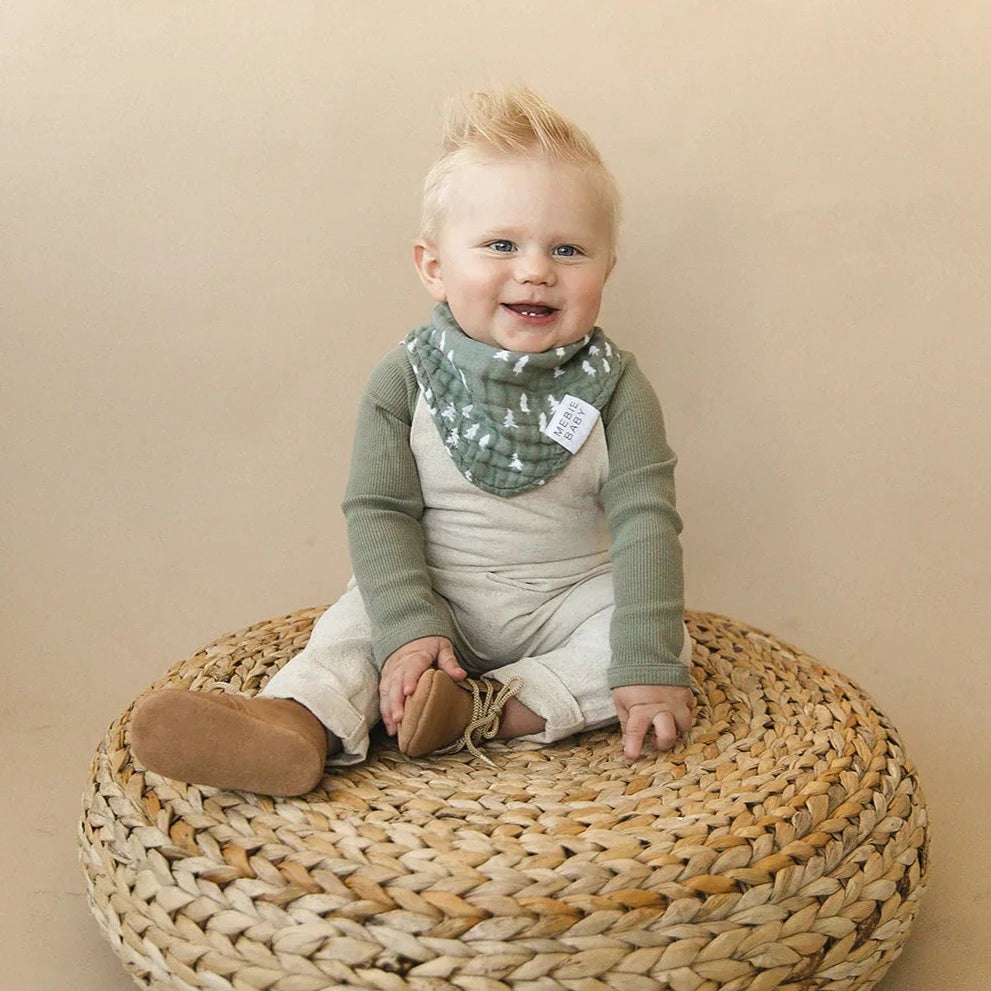 Cream wall with a rattan poof, and a little boy sitting on it wearing the Pines Bib by Mebie Baby.