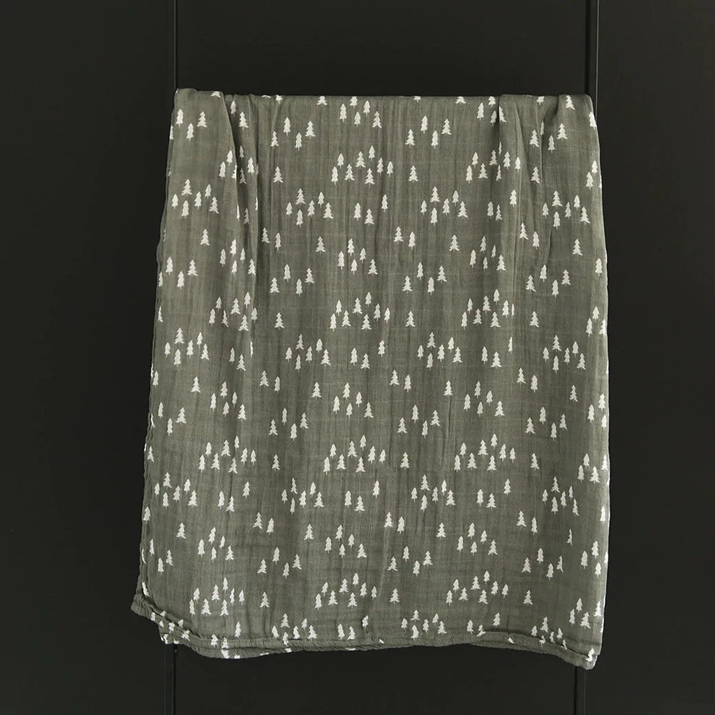 Dark wall with a black metal blanket ladder, and the Pines Muslin Swaddle by Mebie Baby hanging over it. Swaddle is an olive green colour with white pine trees.