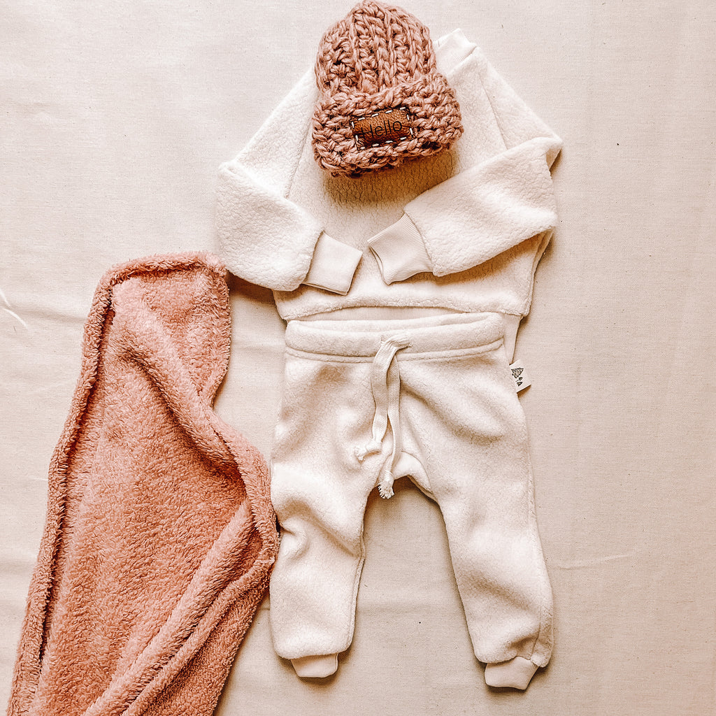 Polar Bear crew and pant by Petit Nordique, paired with a pink 'Hello' Take Me Home beanie by Petit Nordique, as well as a pink sherpa blanket by Petit Nordique. Laid on a flat beige surface. 