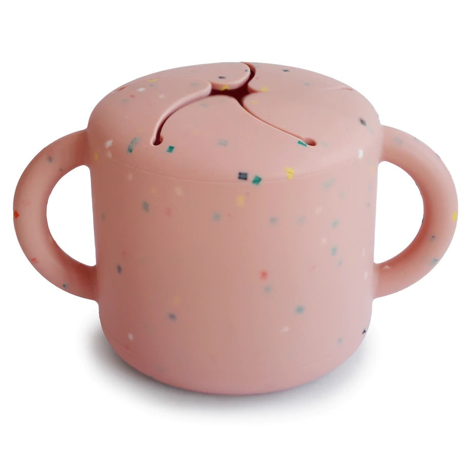 White background with Silicone Snack Cup in Powder Pink Confetti by Mushie. Snack cup has 2 handles, and a spill proof top, in a light pink with sprinkled confetti.