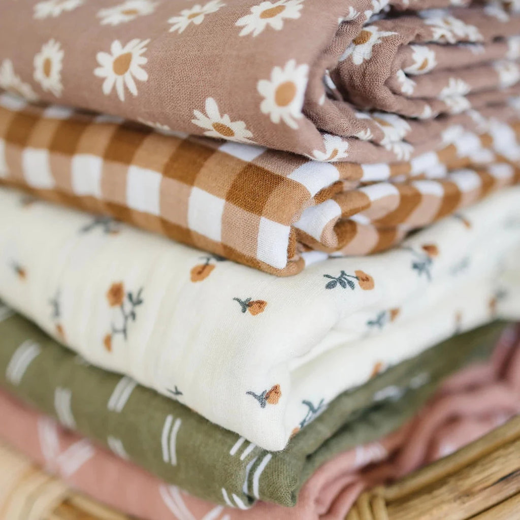 Close up of a stack of Muslin Quilts by Mebie Baby, including Daisy Dream, Gingham, Cream Floral, Olive Strokes, and Just Peachy.