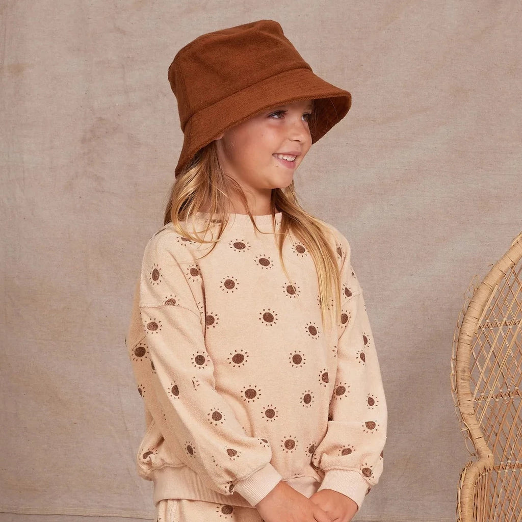 Model wearing Wide Brim Bucket Hat in Chocolate by Rylee and Cru. Model is also wearing Rylee and Cru apparel in a natural colour with suns. Rattan chair in background, all in front of a beige wall. 