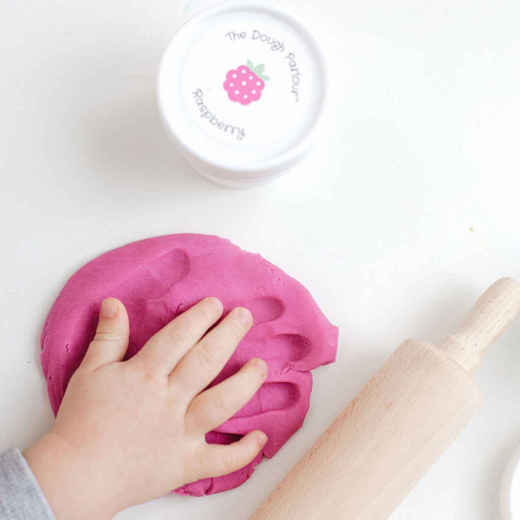 White table with an unopened container of Raspberry Play Dough by Dough Parlour, and a hand playing with play dough. Raspberry play dough is a fuchsia colour.