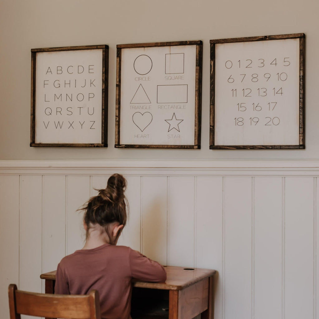 White wall with a little girl sitting at a desk, and the Numbers/Letters/Shapes Wood Sign Set by Restored Signs & Decor hanging on the wall. Comes with 3 signs, 1 is the alphabet, the other is different shapes, and the last is numbers 0-20. All signs are white, with black text, and wooden borders.