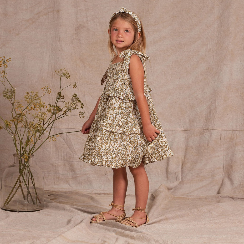 Model wearing Ruffled Swing Dress in Golden Ditsy by Rylee and Cru. and wearing a matching headband. Vase in background with green and yellow flowers, with a beige backdrop. 