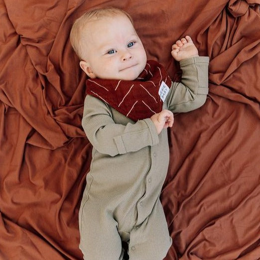 Overhead view of baby laying down on rust blanket, wearing a Rust Mudcloth Bib by Mebie Baby. Triangle bib is rust, with white lines all over.