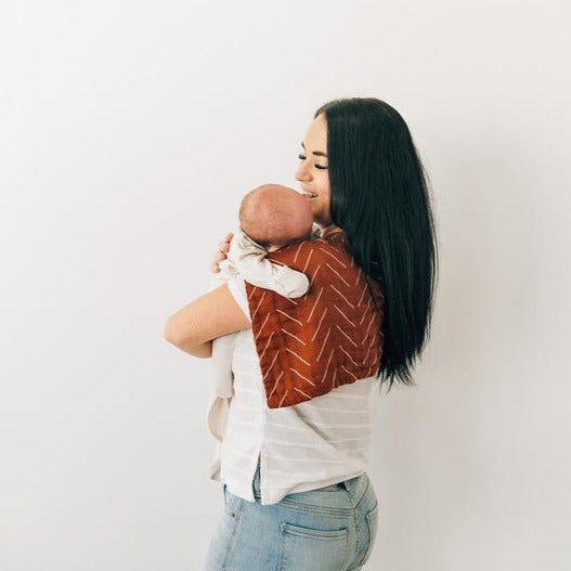 White background with mama holding a baby, back to the camera, and a Rust Mudcloth Burp Cloth by Mebie Baby draped over her shoulder. Burp cloth is rust with white lines.
