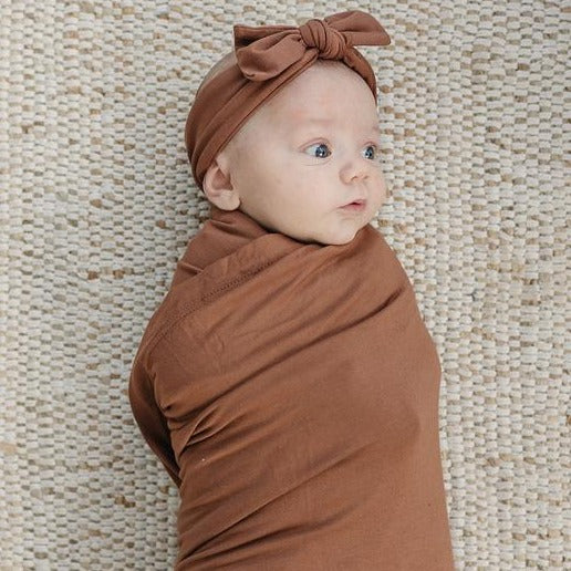 Woven background with a baby wrapped up in a Rust Stretch Swaddle by Mebie Baby. Swaddle is rust, with tons of stretch.
