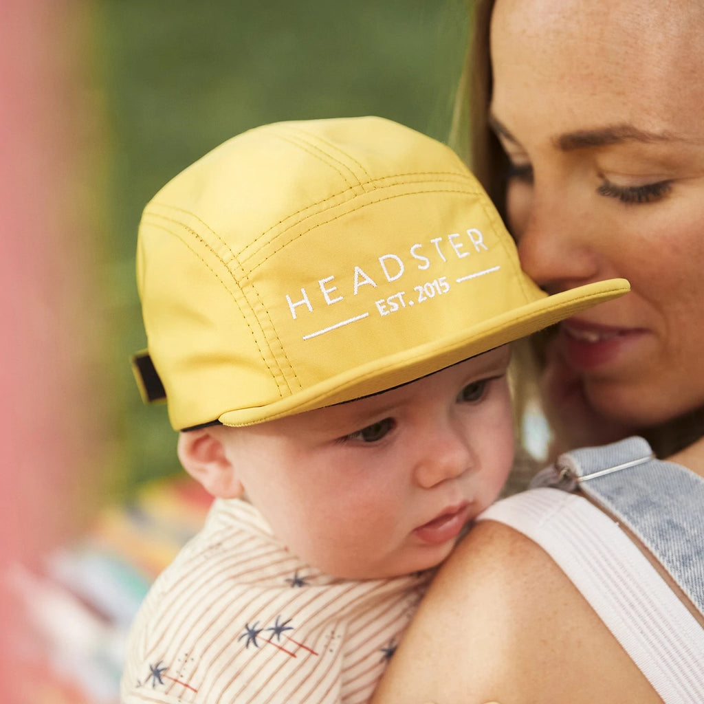 Close up of baby in moms arms wearing a Salty 5 Panel hat in Sunny Day by Headster. 5 Panel hat is a bright yellow with white stitching that says "Headster" across the front.