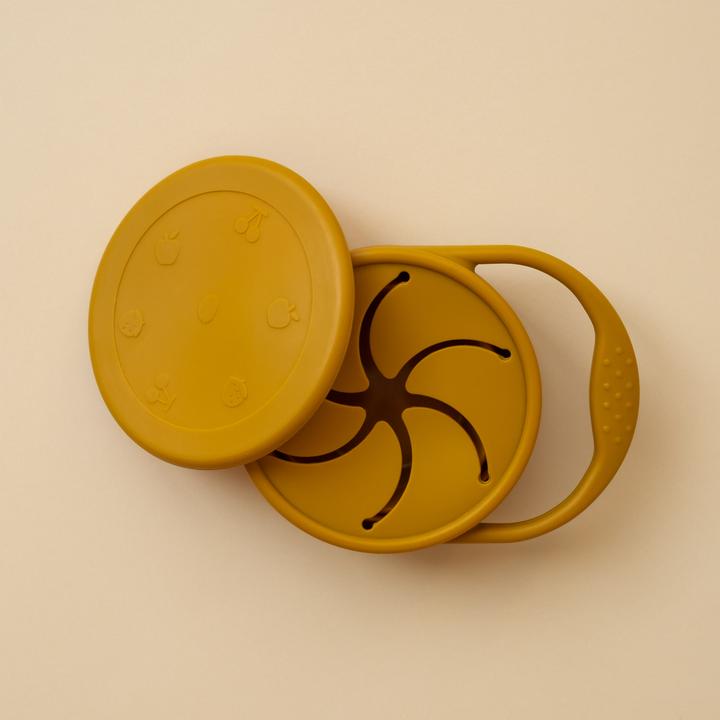 Beige background with a Silicone Snack Cup in Ochre by Minika. Snack cup has a small handle to carry, and a lid as well, all in the colour ochre.