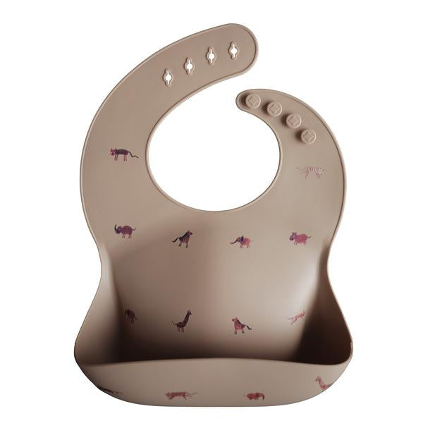 White background with a Silicone Bib in Safari Tan by Mushie. Bib is a tan colour with reddish safari animals, with a deep pocket on the front, and a rounded neck fastener at the back.