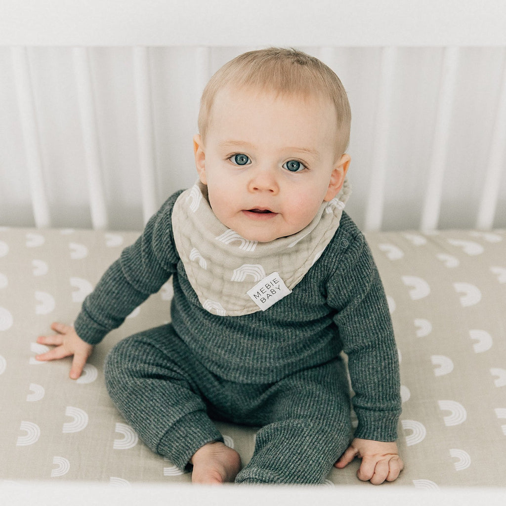 Baby boy sitting up in white crib, wearing a Sand Rainbow Bib by Mebie Baby. Triangle bib is a pale beige/grey with rainbows all over.