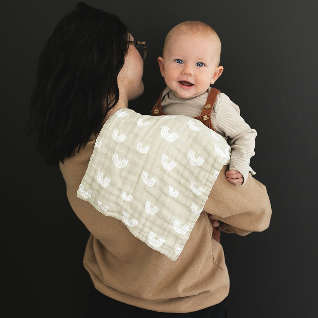 Sand Rainbow burp cloth by Mebie Baby on a mothers shoulder, while she holds her baby. She is standing in front of a black wall. 