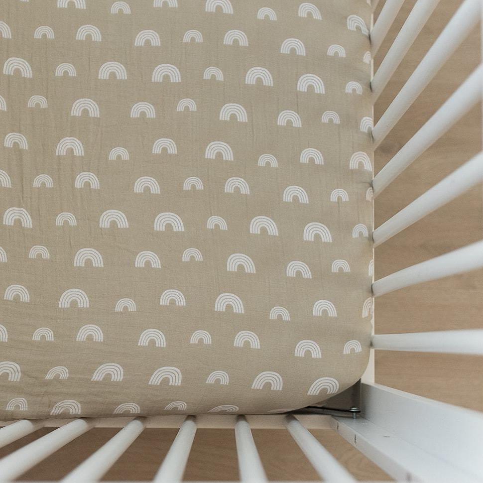 Overhead view of a white crib, and a Sand Rainbow Crib Sheet by Mebie Baby on the mattress. Crib sheet is a beige/grey with white rainbows all over, and fits snug.