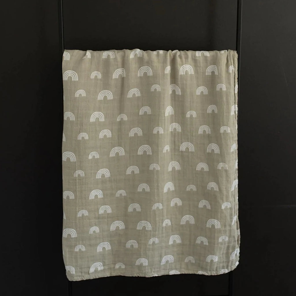 Black wall with a black metal blanket ladder and the Sand Rainbow Muslin Swaddle by Mebie Baby hanging on it. Swaddle is a creamy beige colour with white rainbows all over.