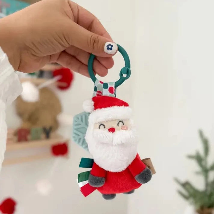 Holiday Santa Itzy Pal™ Plush + Teether being held by a hand in front of a white wall, with a book shelf that is decorated for the holidays and a green plant in the background. 