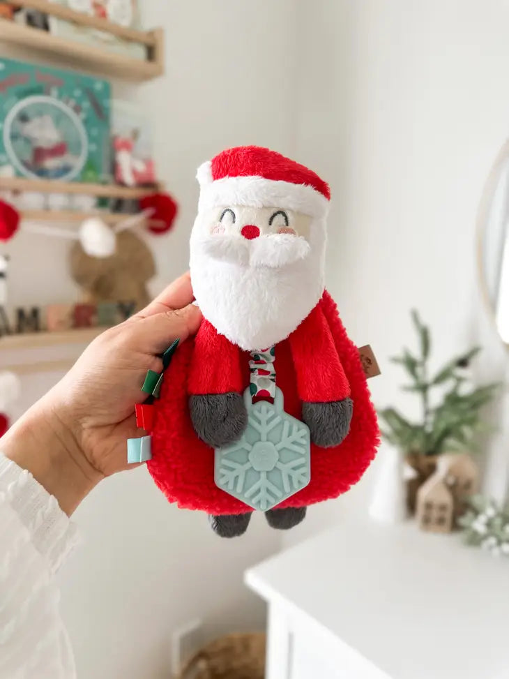 Holiday Santa Itzy Lovey™ Plush + Teether Toy Being held up by a hand in front of a book shelf, and dresser with winter and holiday decor, 