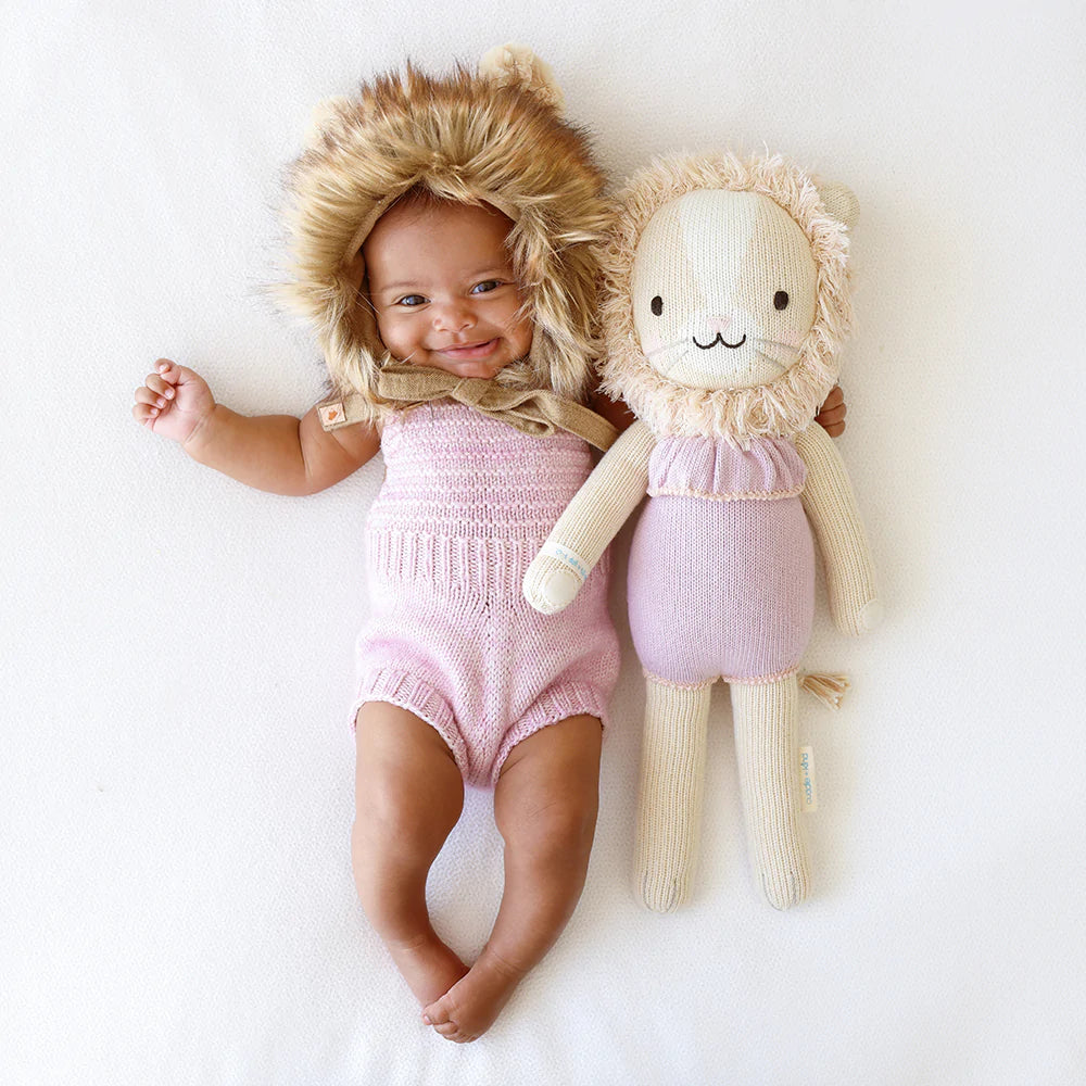 White background with a baby girl laying down, with a lion mane bonnet with a Savannah The Lion by Cuddle and Kind beside her.