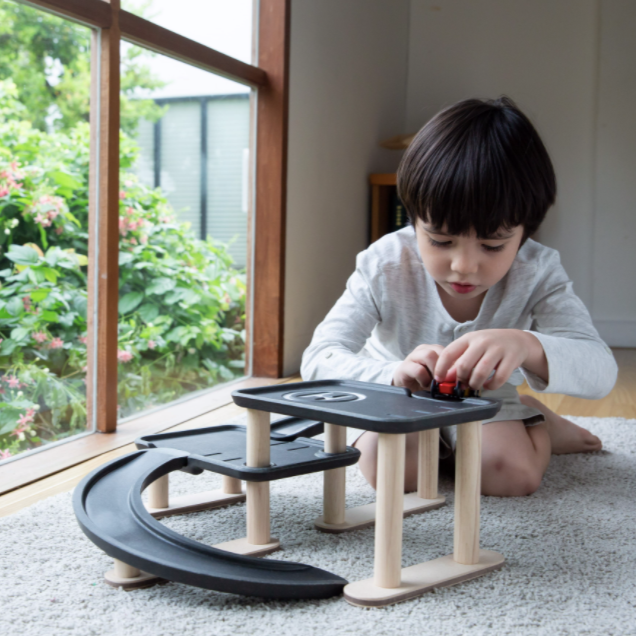 A little kid is bent over in a living room, playing with the Race N Play Parking Garage by PlanToys.
