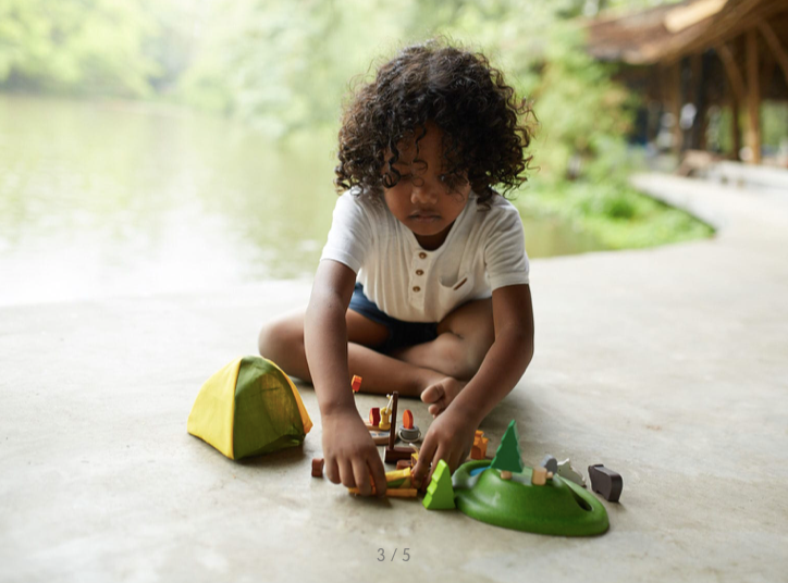 A little kid sitting outside playing beside the water with the Camping Set by PlanToys.