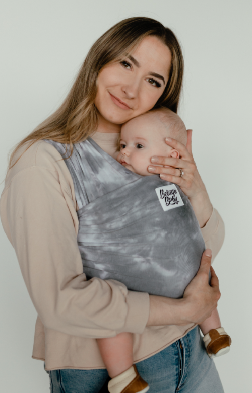 Model wearing Wrap Carrier in The Hendrix in a grey tye dye by Beluga Baby, with a baby in carrier, with a beige background. 