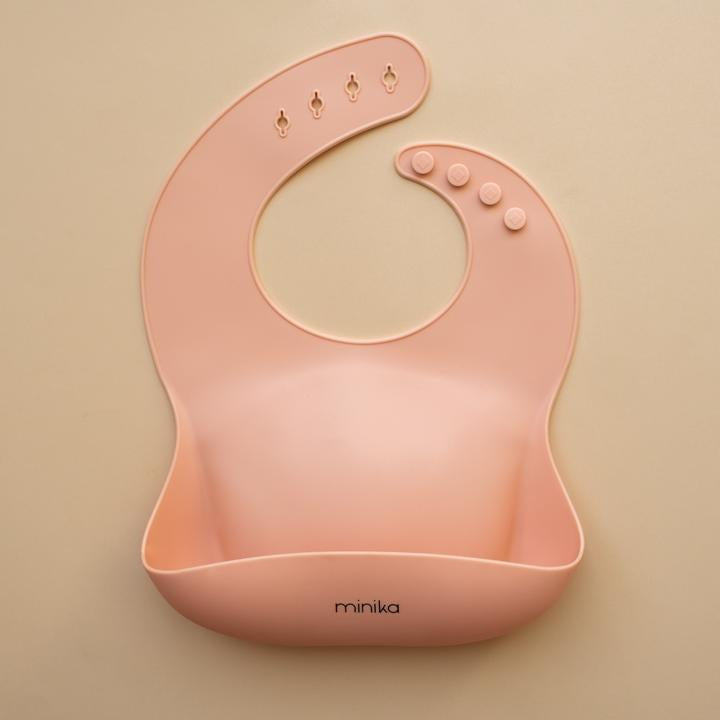 Beige background with a Silicone Bib in Blush by Minika. Bib is blush colour, and has a pouch on the front.