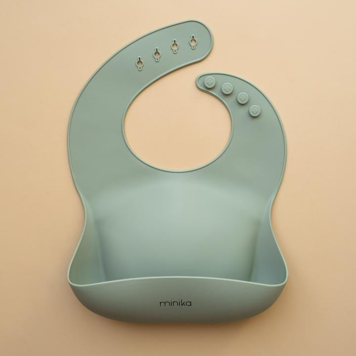 Beige background with a Silicone Bib in Sage by Minika. Bib is sage colour, and has a pouch on the front.
