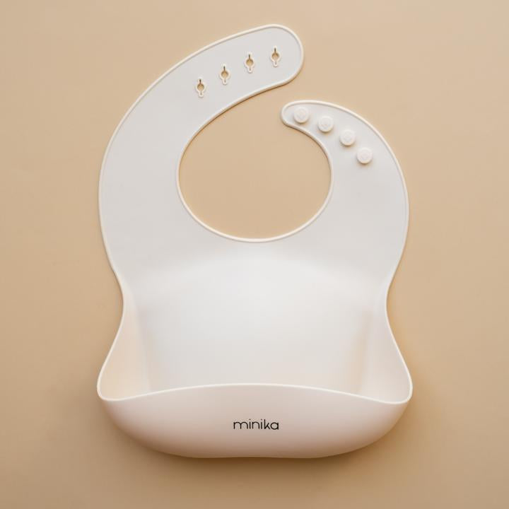 Beige background with a Silicone Bib in Shell by Minika. Bib is white colour, and has a pouch on the front.