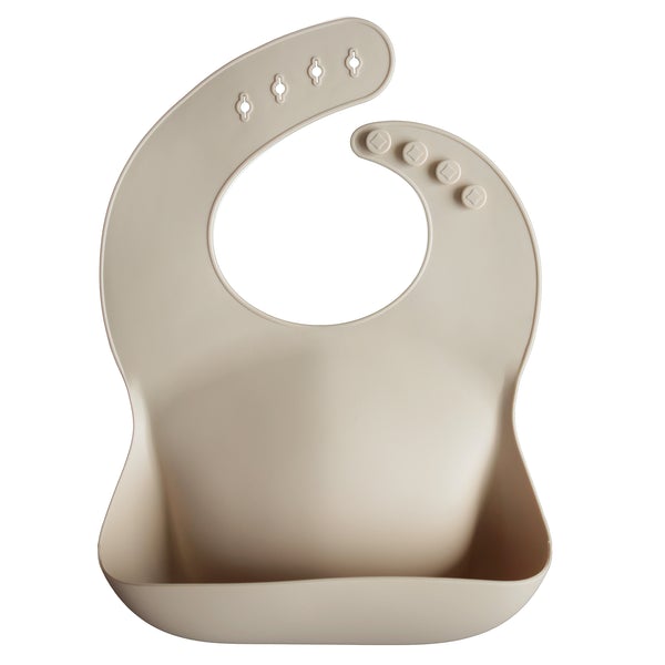 White background with a Silicone Bib in Shifting Sand by Mushie. Bib is a beige colour, with a deep pocket on the front, and a rounded neck fastener at the back.