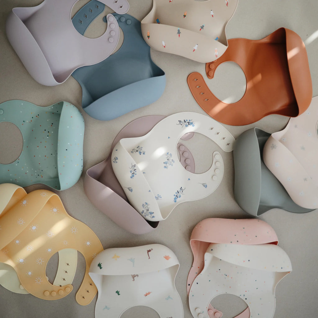 Grey background with an overhead view of a whole pile of Silicone Bibs by Mushie in numerous colours, all stacked on top of eachother, and sprawled out.
