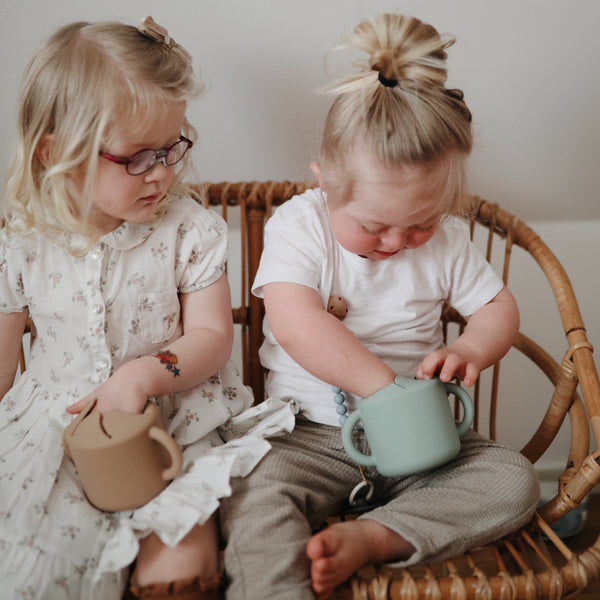 Two little girls sitting on a rattan loveseat, holding Silicone Snack Cups by Mushie.