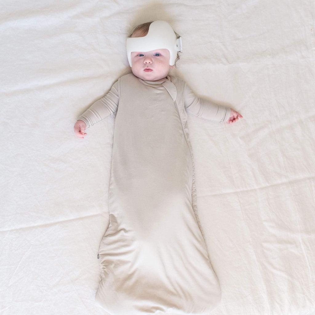 Cream background with overhead view of baby laying down, wearing a Sleep Bag 2.5 Tog in Oat by Kyte Baby. Sleep bag is a neutral/grey colour, with a side zipper going all the way down to the bottom.