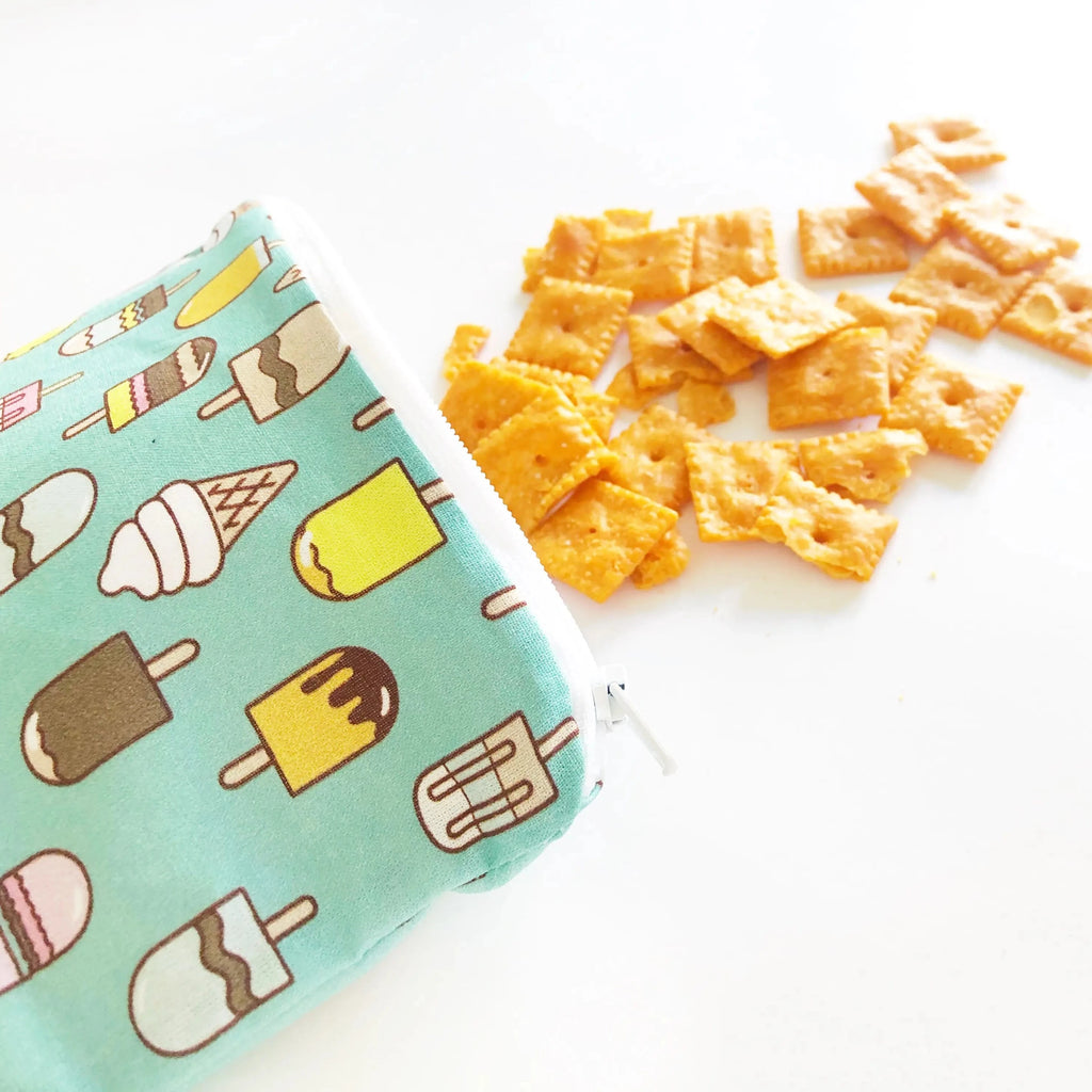 White background with a Resuable Snack & Everything Bag in Ice Cream Social by Itzy RItzy, with cheese crackers coming out of it. The bag is teal with different colour popsicles and ice cream cones.