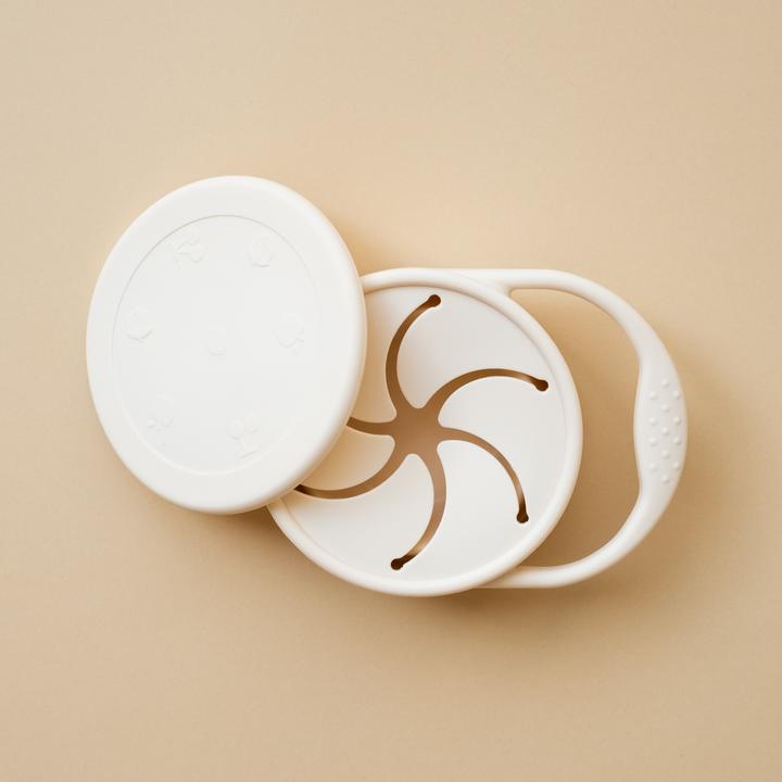 Beige background with a Silicone Snack Cup in Shell by Minika. Snack cup has a small handle to carry, and a lid as well, all in the colour white.