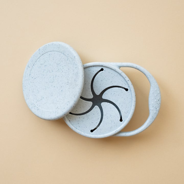 Beige background with a Silicone Snack Cup in Ice by Minika. Snack cup has a small handle to carry, and a lid as well, all in the colour ice blue speckled.