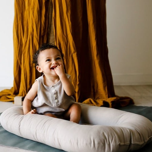 White wall with an ochre blanket hanging, and a little boy sitting in the Snuggle Me Organic Infant Lounger in Birch by Snuggle Me.