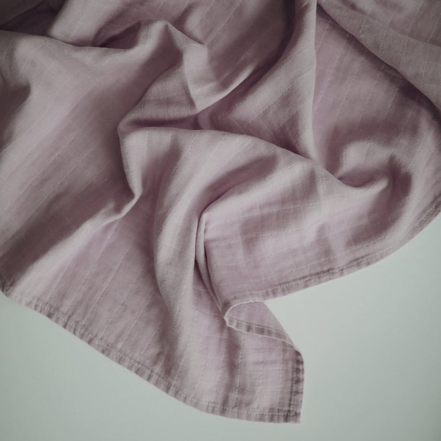 Muslin Swaddle Blanket Organic Cotton in Soft Mauve by Mushie laid on a flat white surface. 