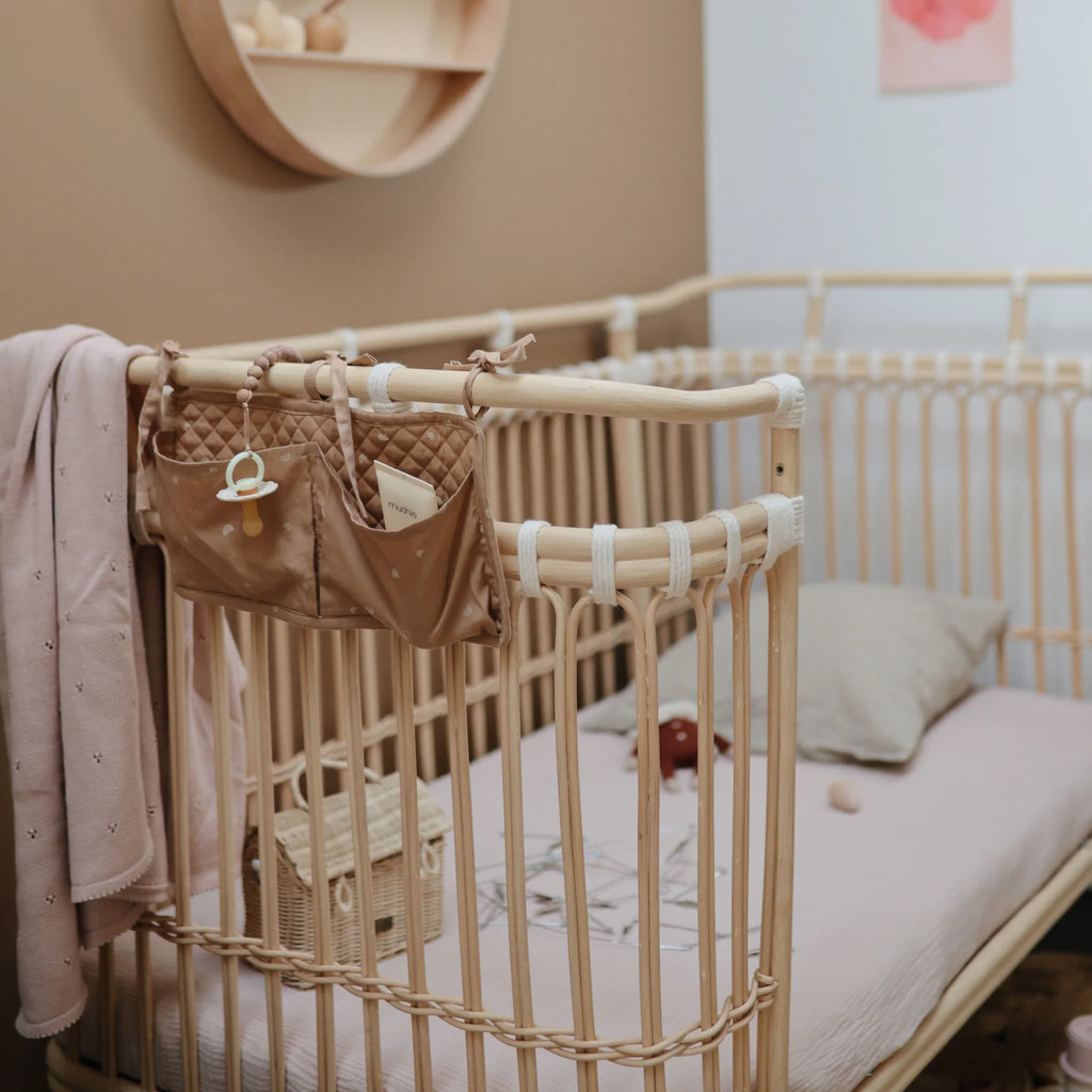 Close up of a rattan crib in a nursery, with the Crib Storage Pocket in Rainbows by Mushie attached to the side. The storage pocket has a soother, and baby lotion in it.