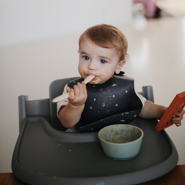 Baby sitting at high chair eating, using a Silicone Suction Bowl by Mushie.