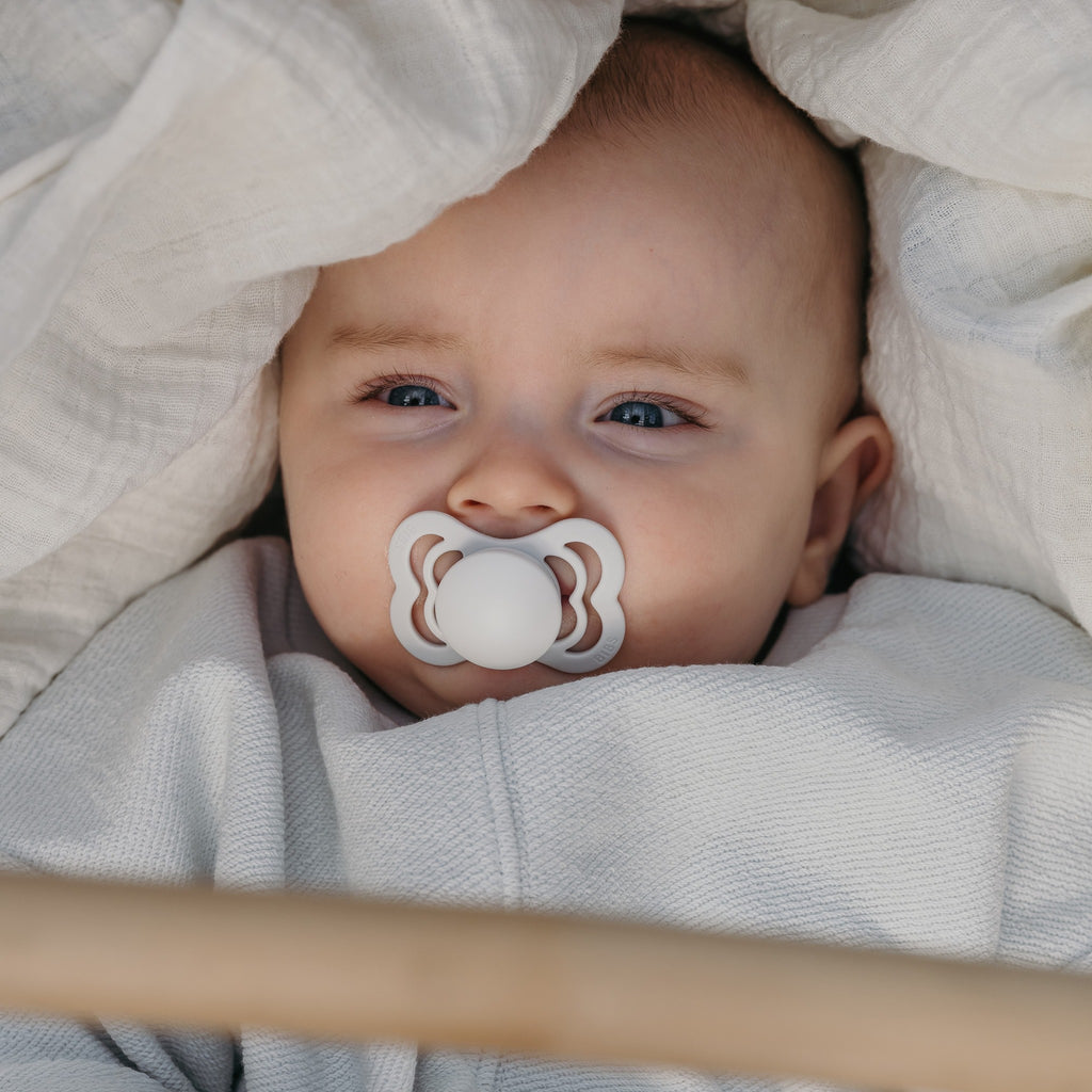 Close up of baby laying in a stroller, with the Supreme Size 1 Pacifier in Ivory by Bibs in their mouth.