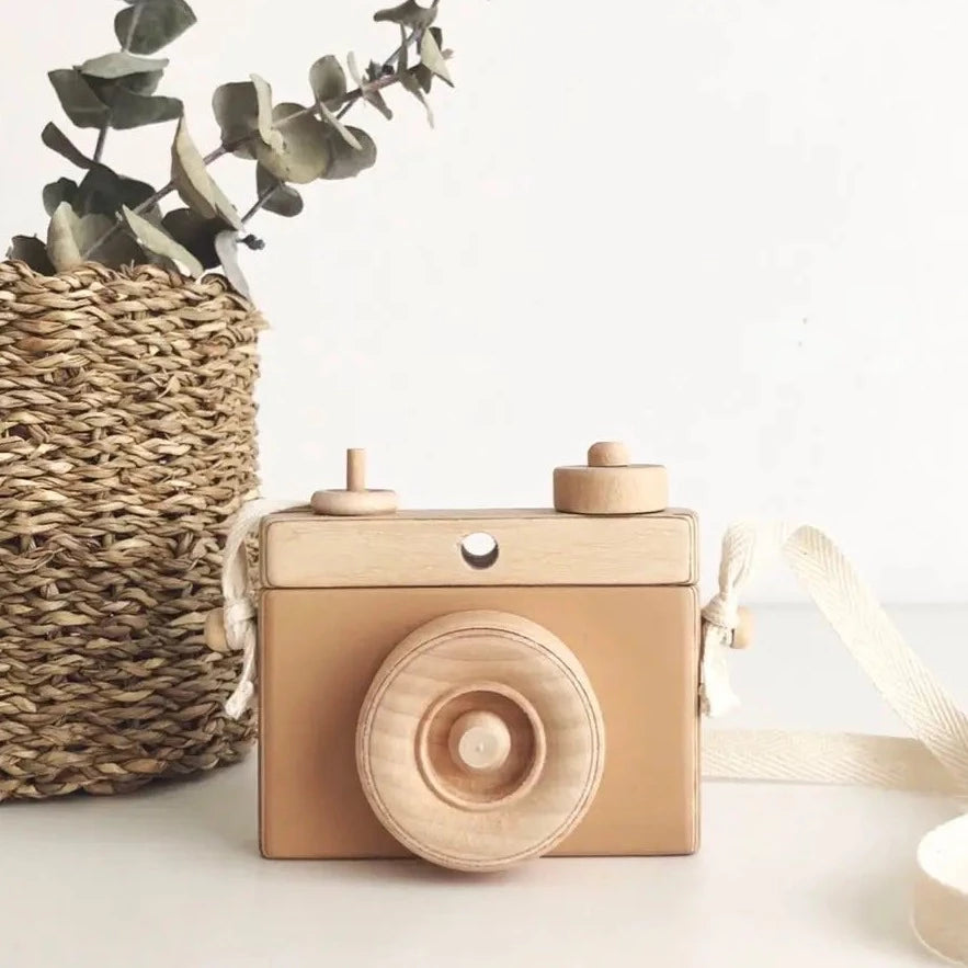 White background with a rattan basket with greenery, and a Wooden Camera in Tan by Little Rose & Co. in the middle. Camera is wood with tan accents, and a canvas strap.