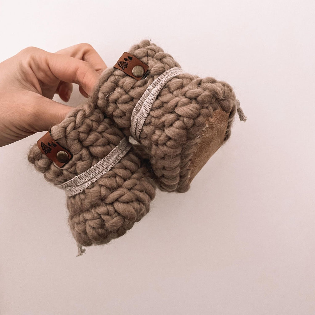 Taupe booties with greige ties and leather soles and tags, by Petit Nordique. Booties being held up by hand in front of a white wall. 