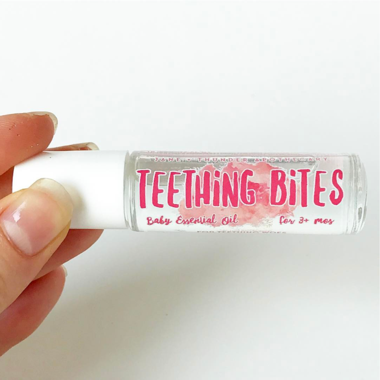 White background with hand holding Teething Bites Baby Safe Essential Oil by Jane + Thunder. Text says "Teething Bites" in pink.