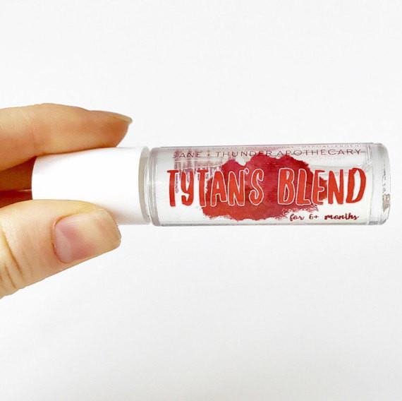 White background with hand holding Tytans Blend Baby Safe Essential Oil by Jane + Thunder. Text says "Tytan's Blend" in red.