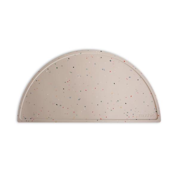 White background with Silicone Place Mat in Vanilla Confetti by Mushie. Placemat is a beige colour with multicolour sprinkles, made of silicone, and in a semicircle.