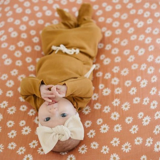 Overhead view of a baby girl laying down on a Daisy Dream swaddle, wearing the Vanilla Organic Cotton Ribbed Head Wrap by Mebie. Head wrap is a cream colour with a large knot bow.